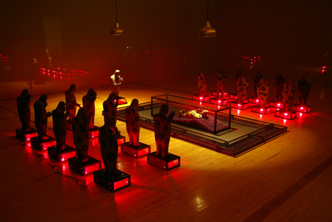 CARLOS RUNCIE TANAKA, "Tiempo detenido / No Olvidar", 1997-2006, Variable dimensions, Installation - 22 ceramic figures, 11 glass/metal cases with red light, 10 glass/metal cases containing red marbles, 1 glass/metal urn containing 1 ceramic figure and three lamps hanging from the ceiling in a room with inner walls covered completely by glass panels. Taking inspiration from his hostage experience at the Japanese Ambassador’s Residence where he was held captive with many others by the MRTA (Movimiento Revolucionario Túpac Amaru) in Lima, December 1996, the artist creates this installation with allusions to violence and the lack of communication. Time detained – Do not forget